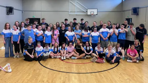 Members of the group line up with three rows, the front sitting and the back two standing. Many members wear club colours of blue and white. Others, volunteers it appears, wear black tops with blue trim. They are in a sports hall with wooden floors and basketball nets around the walls.