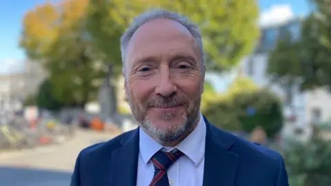 A man called Paul Montague. He is wearing a navy suit with a white shirt and blue and maroon stripey tie. He has a short greying beard and is stood in front of several trees.