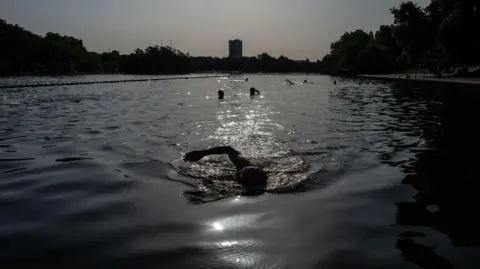 Getty Images People enjoy an early morning swim at the Serpentine Lido. The sun is reflecting off the water, and there are trees lining the banks on either side.