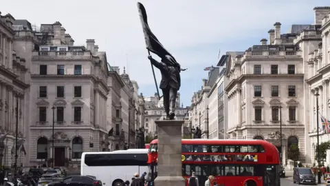 Statue of a man holding a flag in central London. A London bus and people walking around in the background