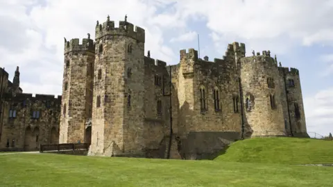 An exterior view of Alnwick Castle in Northumberland. A grey brick, turreted castle with long windows and archways.