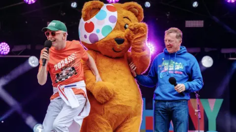 CarFest People on a brightly lit outdoor stage alongside a large orange teddy bear mascot wearing a colorful polka-dot eye patch. Two presenters hold microphones while purple stage lights and event signage are visible in the background.