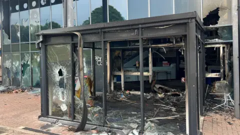 Larne Leisure Centre entrance extremely damaged following rioting. The windows are smashed, there is fire damage and there is a lot of debris on the ground.