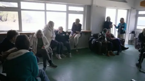 BBC A picture of a group of people sat in a large room located on beach. Some people are stood up, with a majority of people sat down.