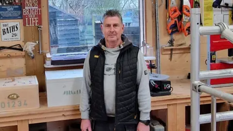 Donna Talbot Maintenance manager John Baker in grey hoody and black gilet in front of a repair window at the Emmaus Cambridge workshop