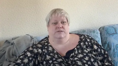 A woman with short blonde hair wearing a long sleeve black floral top sat on a ble floral pattern sofa.