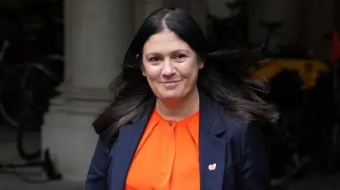 PA Media Nandy has long dark hair and is wearing an orange blouse and navy jacket with a poppy pin on the lapel. She is walking outside past a grey stone column.