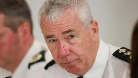 PA Media PSNI chief Jon Boutcher looking at camera. He is in uniform and a colleague appears in the backgorund to his right