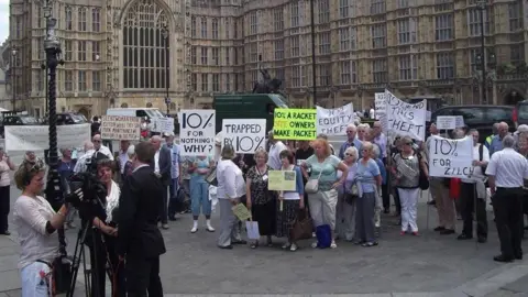 Alan Baldwin A group of park home residents are at Westminster in 2017 holding protest signs about a 10% commission fee. Several people hold placards reading “10% for nothing why?”, “Trapped by 10%” and “10% equity theft”, while a television crew films nearby in front of the building.