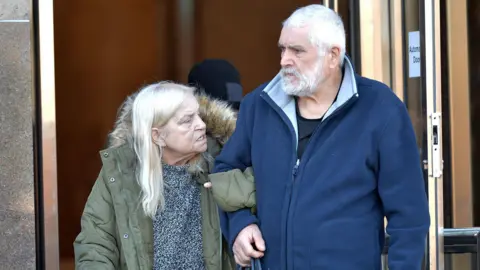 Barbara and Douglas Daniel leaving Glasgow Sheriff Court. Barbara is on the left and is wearing a large green jacket over a grey top. She has white hair. Douglas is on the right and has white hair and a beard. He is wearing a blue fleece.