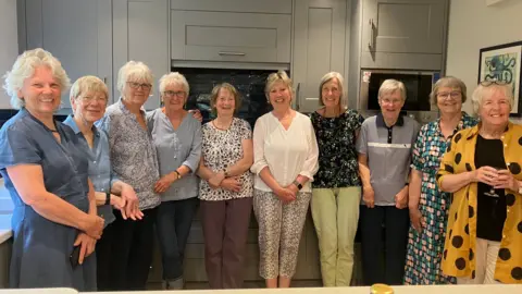 A group of 10 women in their 70s and 80s standing in a large kitchen. They are all smiling.
