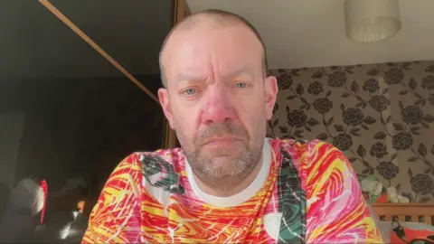 A man with very tightly cropped hair and stubbly beard is wearing a top of swirling red, yellow and pink colours. He is sitting in a bedroom on a video call.
