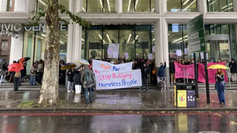 HASL Protesters gathered outside Lambeth Civic Centre holding umbrellas and banners, including one reading “Stop Bullying Homeless People”, during a demonstration in wet weather.