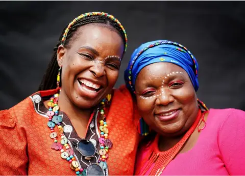 Brian Lawless/PA Wire Tina Sekete (left) and Jane Dlamini smiling. They are wearing bright colours and have white make up on their face.