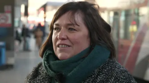 Rosie Trew is seen wearing a dark coat and green scarf is at a bus station platform, with buses and people visible behind her.