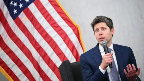 The OpenAI CEO Sam Altman is wearing a dark blue suit with a white shirt and blue and white tie. He is holding and speaking into a microphone while sitting in front of the American flag