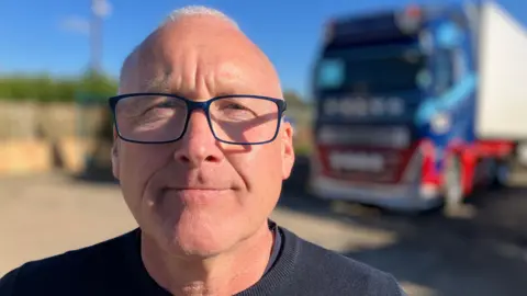 Andrew Sinclair/BBC Haulier Trevor Rowell standing next to one of his trucks.