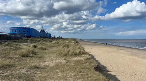 Qays Najm/BBC The beach at South Beach parade in Great Yarmouth. Dunes lead from the promenade onto the sandy beach. There are clouds in the blue sky and people walking along the beach.