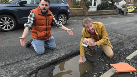 Councillor Andrew Tromans Councillor Andrew Tromans (left) is knelt beside the big pothole that is filled with water. He wears blue jeans, an orange body warmer and checked shirt underneath. David Sheppard (right) is on one knee with his arm inside the water filled pothole, he wears a mustard jacket and beige trousers.