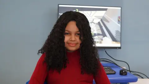 BBC Rama, a 10-year-old girl with long dark curly hair, is wearing a red top and sitting in front of a computer screen showing the video game Minecraft.