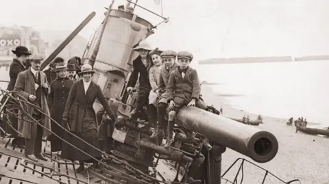 Getty Images A black-and-white photo from 1919 shows three children and a woman sitting on the gun barrel of the submarine. To the left, a crowd of men and women also smile at the camera. In the background, the shoreline of Hastings Beach can be seen. 