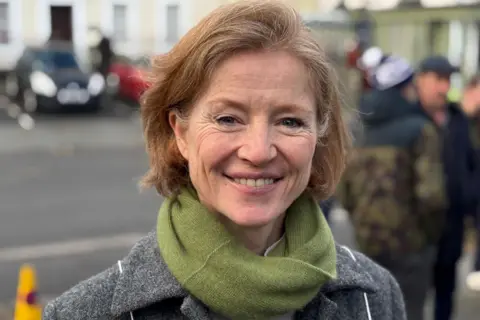 BBC/Mark McAlindon A head-and-shoulders picture of Sarah Dunning - a woman with red hair in a short bob. She is smiling widely and is wearing a green scarf and grey coat. She is standing in front of businesses, cars and a crowd of people.