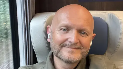 Darryl Matthews Darryl Matthews smiles as he poses for a selfie while sitting on a train seat.