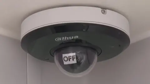 A ceiling-mounted internal CCTV camera. In front of its lens is an automatically movable sign that says "Off".
