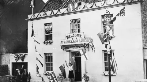 G. Holt, US Army Signal Corps Guernsey woman standing outside house with a sign saying "welcome, at long last liberty"