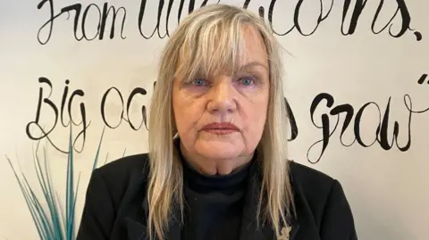 Jean Taylor has long blonde hair and is wearing a black jacket over a black jumper. She is standing in front of a white wall and a grass plant.