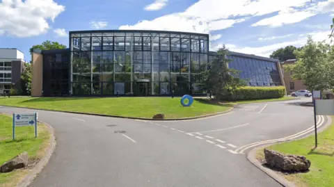 Google A glass building with lots of large plants behind the glass. It is at the junction of several roads, with a grassy space in front.