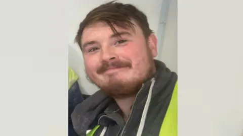 Dean Green is smiling at the camera. He has brown eyes, brown hair and a brown beard and moustache. He is wearing a high-viz jacket over a black hoodie.