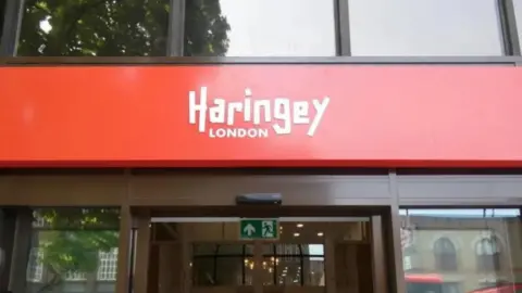 A red sign above a council building entrance reads "Haringey London" written in white text. Below a green fire exit sign hangs in the doorway. 