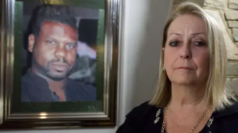 Carole Charles, a middle-aged woman looking sad. A photo of her husband is visible on the wall behind her. She is made up smartly and wears a black top and a gold-coloured chain. She has blonde hair coming down to her shoulders.