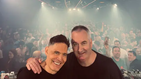Two men in black T shirts, arms around one another, stand in front of a busy crowd