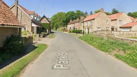 Google A view of a street looking up a slight incline. It is lined by small grass verges and there are cottages either side. The weather is sunny.