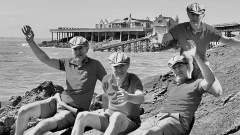 #nicolaexplores Four men in flat caps raising their hands on the coast with Birnbeck Pier in the background