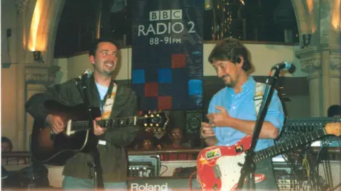 Paul Casey Musicians Paul Casey and Chris Rea share a BBC Radio 2 stage. A promo for radio 2 can be seen at the back of the stage, between Casey, who is on the left and wearing a green jacket, and Rea on the right, and in a blue shirt