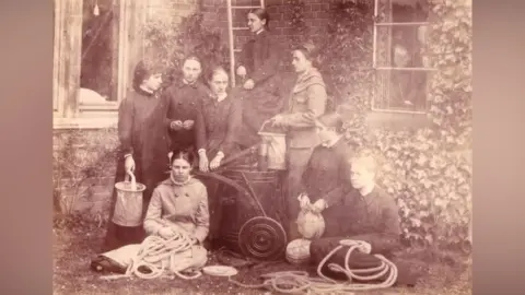 Courtesy of The Mistress and Fellows, Girton College, Cambridge A sepia-toned black and white image of eight women in the Girton College fire brigade, outside and against the corner of an ivy-clad building. They are surrounded by fire equipment. Two seated women are holding ropes, there is a pump in the middle, a third seated woman is holding a hat. Two standing women are holiding buckets and one of the women is perched on a ladder leaning on the wall 