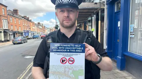 A uniformed police officer stands on a town-centre pavement holding a poster about new powers to tackle unacceptable behaviour in the area. The poster includes symbols related to alcohol and antisocial behaviour and a map of the affected zone. A row of brick-fronted shops and parked cars lines the street behind.