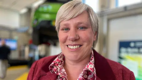 Emma Baugh/BBC Ms Nicholls is smiling at the camera while wearing a red blazer and red flower shirt. She has short blonde hair and blue eyes.