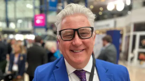 Kevin Bentley with thick grey hair and big black glasses smiling directly at the camera. He is wearing a bright blue suit. Out of focus behind him is a party conference.