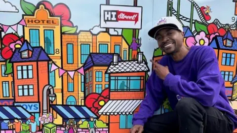 Artist Marvin Mudzongo, in a purple jumper and black jeans, squats next to a colourful digital picture of buildings. 
