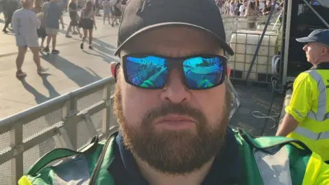 Handout Steve Alcock, who has a ginger beard and is wearing a baseball cap, reflective sunglasses and a green medical uniform, poses for a selfie with the On The Waterfront stage in the background.