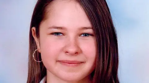 Professional portrait photo close up of Michelle Stewart - a young girl with blue eyes and long brown hair, looking into the camera and smiling. Wearing gold hoop earrings and sitting in front of a blue backdrop