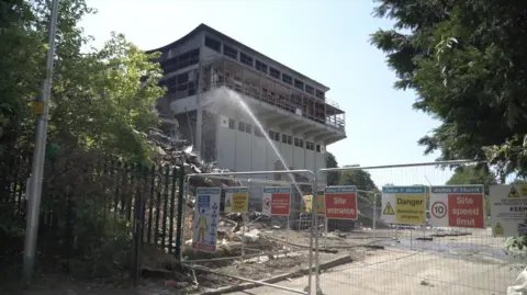The former St John Fisher School in Chatham during demolition, with sections of the building removed, safety fencing in place and water being sprayed to reduce dust.