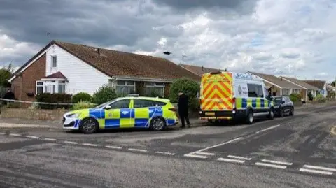 a police van and a police car park either side of a sign saying Holly Road, in front of a modern bungalow.