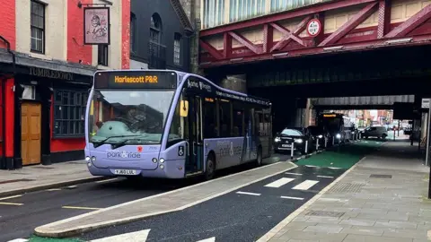 A single lane road with a bus stop in the middle. A Harlescott Park & Ride bus is waiting there, with a line of traffic behind it. A 1.1m-wide pavement "island" is next to the bus. To the right of that is a cycle lane, which is coloured green, apart from a section with a small, two-striped section of zebra crossing