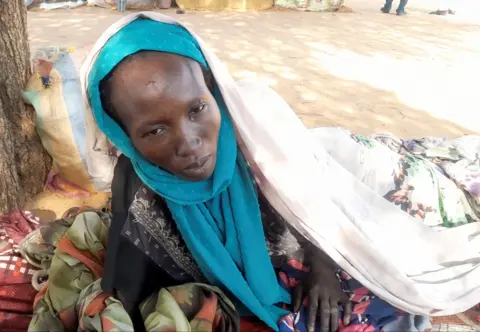 Hamida Adam Ali, in black clothes and a blue shawl is on the floor, with a blanket wrapped over