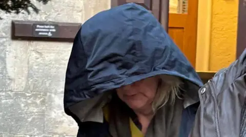 A woman in her 60s emerges from court, attempting to conceal her face by looking down with the navy hood of her coat hanging over her head. She has shoulder length blonde hair and a straight face. The shoulder of another person walking alongside her wearing a brown coat can be seen. The door to the court, a stone building, is behind her.